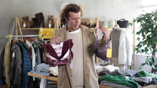 Focused male customer holding bras in hands and selecting underwear in modern clothing store 