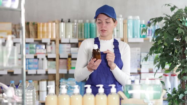 Worker woman in overall buyer select expectorant syrup medicine, reads composition. Client pick up remedy for treatment disease. Client check for absence of harmful substances content 