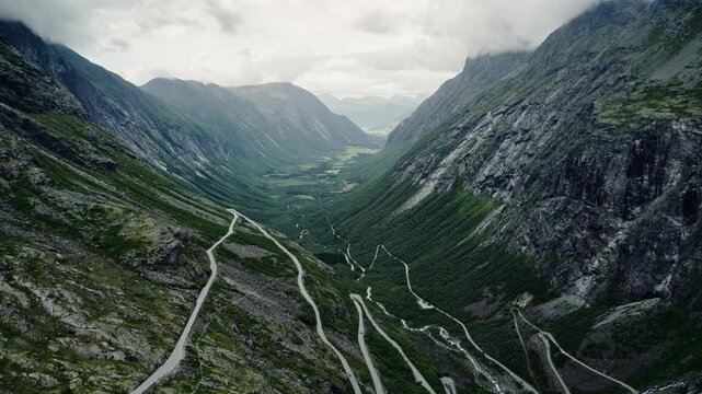 Cinematic drone pull back shot revealing the winding Trollstigen road and Isterdalen valley. View of the visitor center and rugged mountains in More og Romsdal, Norway, under a moody sky.
