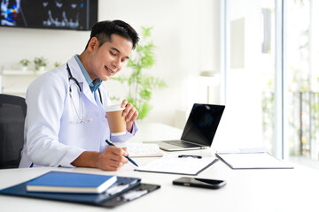 Fototapeta premium Doctor working in office, holding a hot coffee drink cup, reading medical document, focused therapist physician wearing gown sitting at desk in hospital, analyzing patient diagnosis or report.