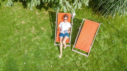 Fototapeta premium Young girl relaxes and reading book in summer garden in sunbed deckchair on grass, relaxing outdoors in green park on weekend, aerial drone view from above