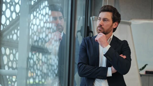 Professional business man stands thoughtfully by modern office window, contemplating complex ideas. Scene evokes feelings of reflection, decision making, strategic planning, deep concentration.