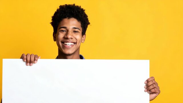 Smiling young man holding a blank white sign with hands against a vibrant yellow background