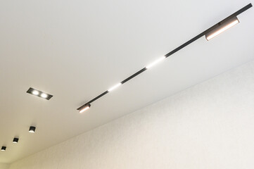 Fototapeta premium A modern ceiling features a sleek black linear track light with adjustable spotlights, complemented by recessed square lights; set against a clean white ceiling and textured wall.