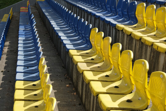 Yellow and blue dirty plastic seats in a forgotten stadium tier. Seats are covered in dirt and dust. Sun bleaching and lack of care are evident on unused rows.