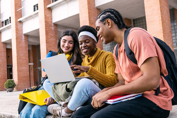 Group of diverse university students smiling and collaborating using a laptop outdoors on campus,...