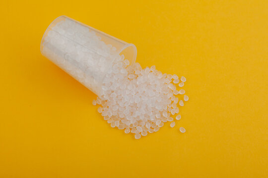 Polypropylene homopolymer (PP-H) granules spilled out of plastic cup on yellow surface. Base polymer for production of plastic packaging. Polypropylene homopolymer resin pellets, close-up