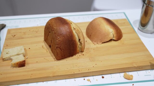 Close up of a freshly baked homemade bread loaf with golden brown crust resting on a wooden cutting board, showing crumbs and soft interior texture in a kitchen environment.