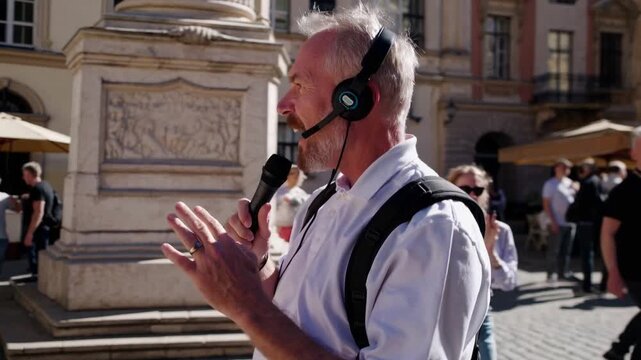 white man tour guide speaking microphone in sunlit historic city square; animated narration about monument details, gestures toward statue, engaging tourists with humor, backpacks and cameras,