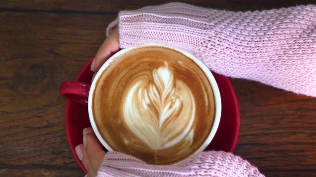 Top down view of a woman's hands in a cozy pink sweater gently holding a red mug of freshly brewed cappuccino with delicate rosetta latte art, resting on a rustic dark wooden table