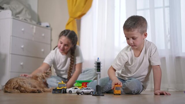 Boy lifts toy traffic light above line of cars and trucks. Puppy rests near sibling girl on floor. Imaginative child continues building. Toy, boy, sibling, toddler act in quiet coordination.