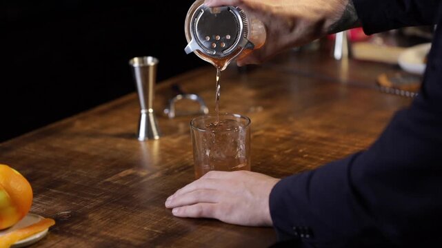 Bartender serving an old fashioned cocktail with a strainer into a glass with a big ice rock.
