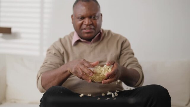 Sport fan watching TV and throwing popcorn when soccer team get goal, portrait. African american mature man relaxing in front of television set in living room in evening or in weekend, entertainment
