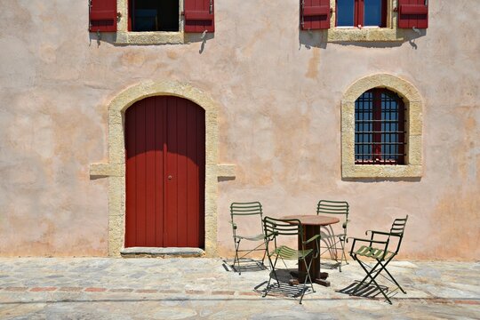 Rural landscape with scenic view of a local gallery facade with a Venetian stucco wall, red wooden arched door and bistro patio furniture in Kapsali, Kythira island Attica, Greece.