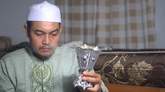 A religious Muslim man wearing a white kufi and green koko shirt holding a traditional silver incense burner (Bakhoor). He is enjoying the aromatic fragrance and smoke in a cozy room. Concept of Islam