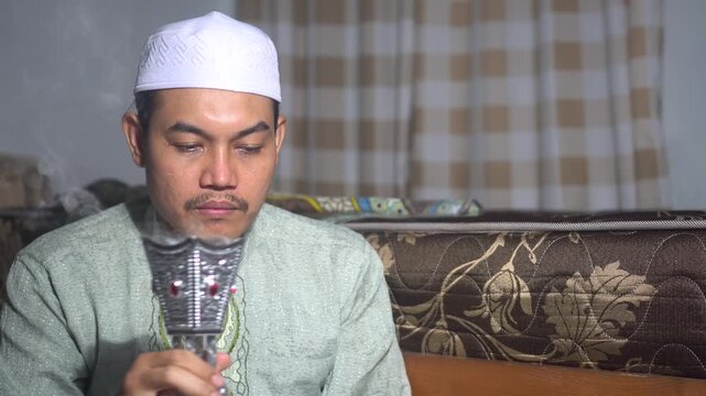 A devout Muslim man in traditional dress and skullcap holding a silver incense burner with aromatic bakhoor smoke. Authentic Middle Eastern and Islamic cultural lifestyle concept for prayer, meditatio