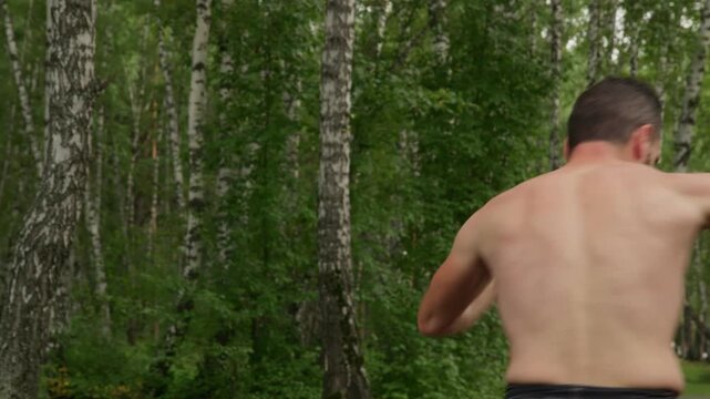 A muscular, shirtless man with a beard practices shadowboxing and martial arts in a beautiful birch tree forest. He trains intensely, showing strength, focus,