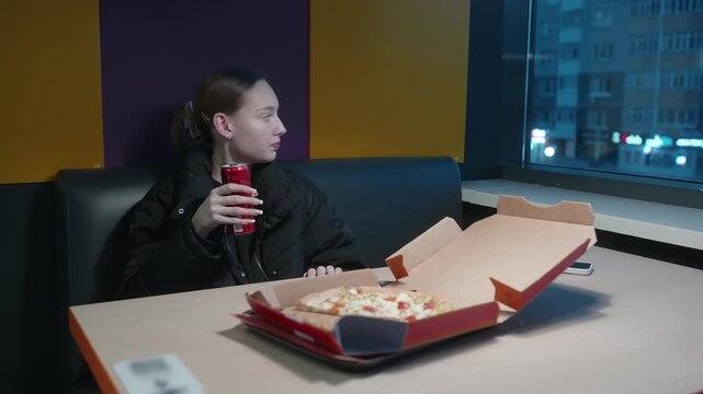 A young woman in a black jacket sits alone in a cafe booth at night, drinking a soda. An open pizza box is on the table in front of her. Urban setting with city lights visible.