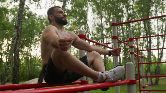 A shirtless, bearded man meditates while sitting on red parallel bars at an outdoor calisthenics park. He practices mindfulness and relaxation after a workout in a green park.