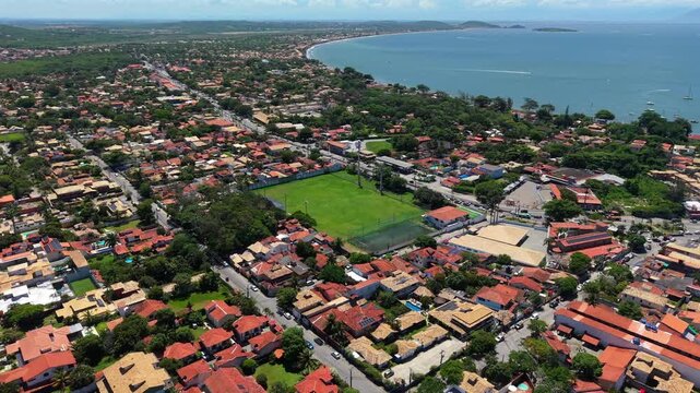 Residential football field approach at Campo da Sociedade Esportiva de Buzios, with ocean and beach visible in the background. Forward descending aerial during clear afternoon in 4K.