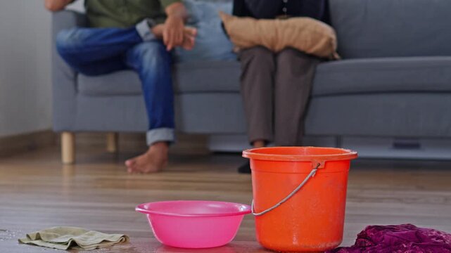 Home roof leak concept showing water dripping into containers while frustrated couple sits on couch