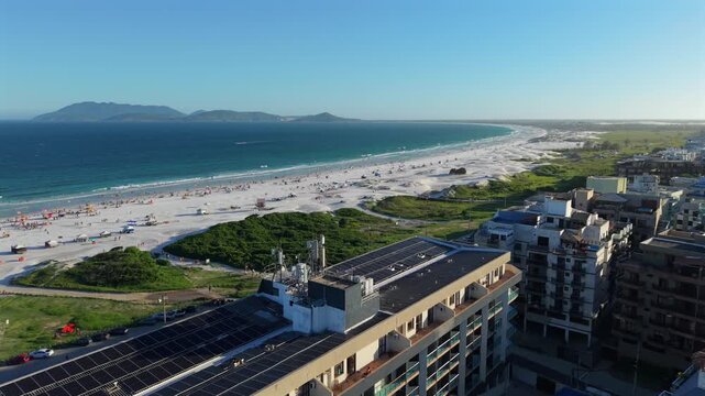 Gliding over shoreline buildings, white sands and bathing tourists at Praia do Forte, Cabo Frio during afternoon, with clear skies and distant horizon. Slow, forward wide-angle aerial in 4K.