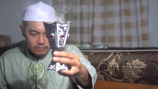 A religious Muslim man in traditional dress and white kufi cap burning aromatic bakhoor incense in a silver burner at home. He is wafting the fragrant smoke towards himself for a spiritual and calming