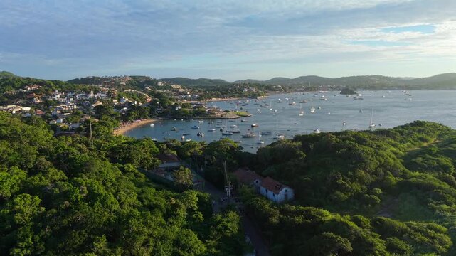 Vegetated hills, beach, and residential area at Praia dos Ossos, Brazil, with multiple boats docked in the ocean during sunset. Forward wide-angle glide.