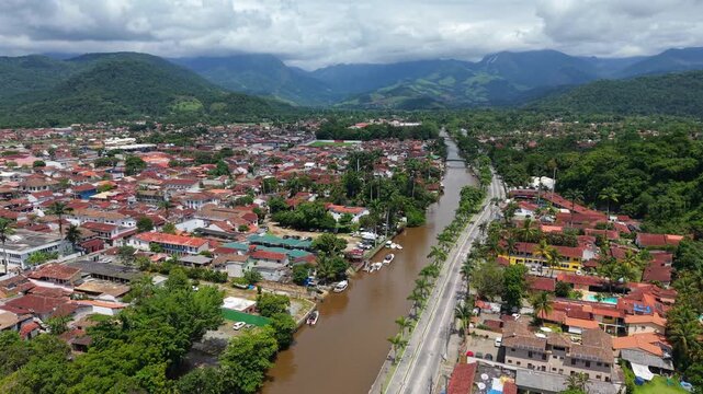 Forward aerial wide-angle glide over historic district of Paraty at Rio de Janeiro. Frame includes river, colonial houses, and dense jungle hills in the background during partly cloudy day.
