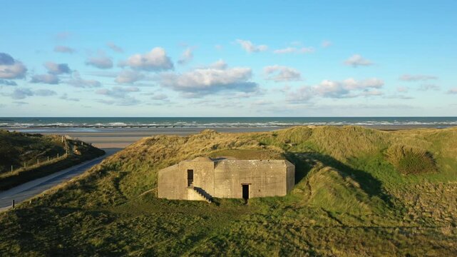 A concrete World War II bunker sits among grassy sand dunes overlooking the shoreline at Utah Beach, Normandy, France, under a blue sky with scattered clouds.