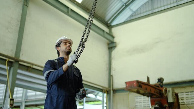 A male factory engineer wearing a safety helmet and uniform operates a heavy duty electric chain hoist with a remote control, ensuring industrial warehouse maintenance and workplace safety standards.