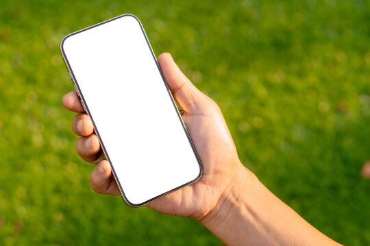 man hand using mockup smartphone at green grass. The phone screen is blank, ready for adding apps, UI, or live sports broadcasts. Close up	