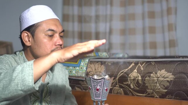 Muslim Asian man in traditional clothes and kopiah using a bakhoor burner at home. Shifting smoke from incense for fragrance and relaxation. Authentic Islamic lifestyle, spiritual tradition, and Middl