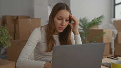 Young hispanic woman at laptop with hand on temple, resting elbow on table, surrounded by cardboard...