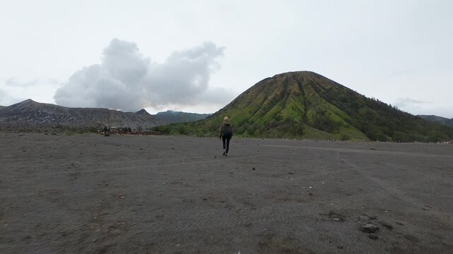 Woman walks alone across black volcanic sand toward lush Mount Batok and smoking Mount Bromo in Indonesia. Scenic travel moment filled with solitude, nature, and adventure.