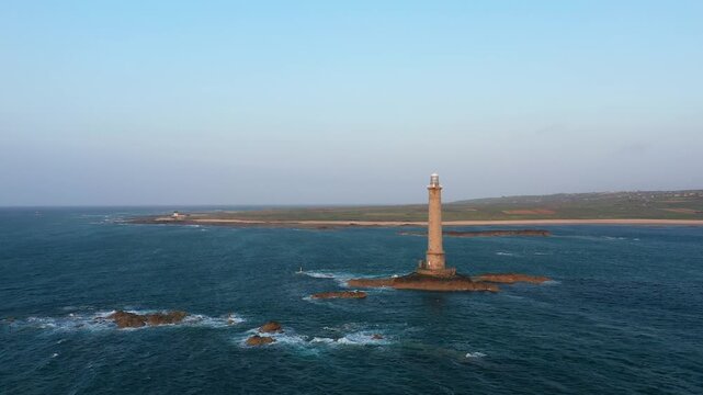 Aerial view of the Phare du Cap de la Hague lighthouse standing on rocky outcrops surrounded by the sea, with the Normandy coastline in the background under a clear sky.