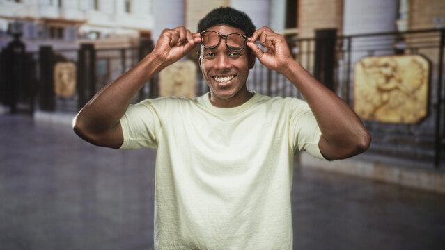 Man adjusts eyeglasses with both hands at temples, smiling broadly in front of ornate railings inside a public building; joyful confidence.