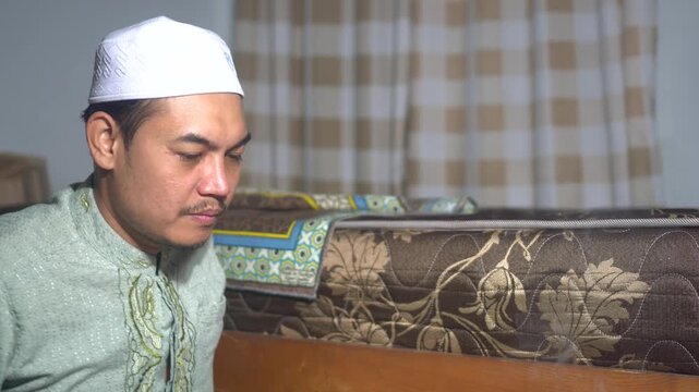 A devoted Muslim man in traditional green batik shirt and white kufi cap performing peaceful prayer and supplication at home. This footage captures spiritual mindfulness, religious devotion, and Islam