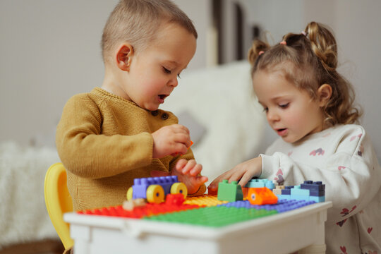 Two young children sit at a table, building with colorful blocks. They focus on their activity, smiling and interacting with each other