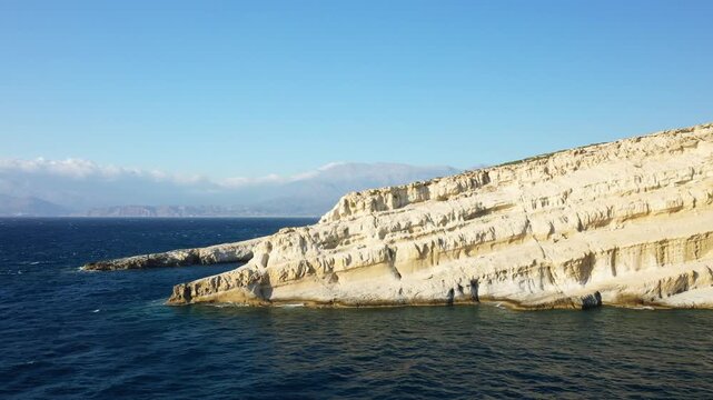 Aerial view of the iconic limestone caves and rugged cliffs along the Matala coast in Crete, Greece, with deep blue sea and distant mountains under a clear sky.