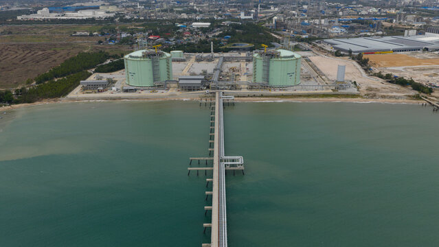 Aerial view of massive green tanks contrasting with the deep blue sea, connected by a long pier stretching into the water, Map Ta Phut, Rayong, Thailand.