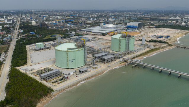 Aerial view of massive LNG storage tanks reflecting the sky's soft light, contrasting with the dark sea and industrial complex, Map Ta Phut, Rayong, Thailand.