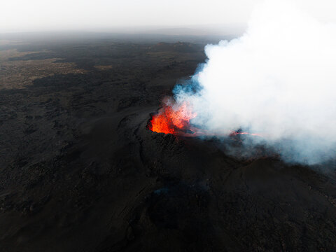 Aerial view of fiery lava erupts, contrasting against the dark volcanic landscape, sending plumes of white smoke skyward, Grindavik, Iceland.