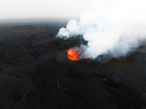 Aerial view of a volcano bursting with fiery orange lava and billowing white smoke, contrasting against the stark black landscape, Grindavik, Iceland.