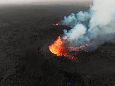 Aerial view of molten lava erupts fiercely, contrasting against the stark, grey volcanic landscape, plumes of smoke billowing into the sky, Grindavik, Iceland.