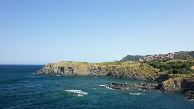 Scenic view of a rugged coastline with clear blue sea and a village on the hillside near Banyuls sur Mer, France. The landscape features rocky cliffs, green vegetation, and tranquil waters under a