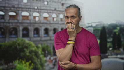 Fototapeta premium Man biting nails with hand to mouth at the roman coliseum building outdoors, long hair and bracelets visible; anxiety contemplation.