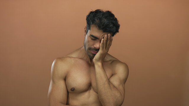 Barechested young hispanic man with hunched posture presses hand to closed eye against brown studio wall; fatigue.