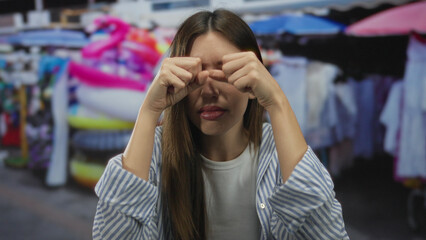 Fototapeta premium Woman rubs eyes at street market booth surrounded by colorful souvenirs and hanging garments; astonishment.