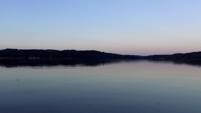 Tranquil view of Lac des Settons at dusk with still water and distant forested hills under a clear sky. Serene natural landscape ideal for relaxation and nature concepts.